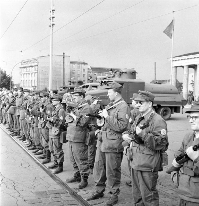 Photographie en noir et blanc. Au premier plan, une longue rangée d'hommes en uniforme, casquette sur la tête et écusson rond sur la manche gauche, se tient au garde-à-vous sur une chaussée pavée traversée par des rails de tramway ; chacun tient contre sa poitrine un pistolet-mitrailleur à chargeur circulaire. Plusieurs portent des lunettes ; ce sont pour la plupart des hommes d'âge mûr. Derrière eux stationnent des camions lance-eau surmontés de tourelles. À droite, les colonnes de la porte de Brandebourg ; un drapeau flotte en haut d'un grand mât. Au fond, des immeubles et des câbles de tramway. Aucune inscription lisible.