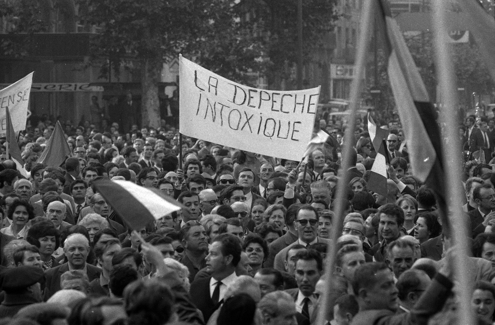 Photographie en noir et blanc, au format horizontal. Une foule dense remplit toute l'image, dans une rue plantée d'arbres et bordée d'immeubles. Au centre, au-dessus des têtes, une banderole blanche tenue par deux hampes porte, écrit à la main en capitales noires : « LA DEPECHE INTOXIQUE ». À gauche, une autre banderole, coupée par le bord de l'image, ne laisse lire que quelques lettres. Plusieurs drapeaux à trois bandes verticales, des drapeaux tricolores selon toute vraisemblance, sont agités au-dessus de la foule, dont un, flou, au premier plan et un grand à droite. Au premier rang, des hommes en costume et cravate, certains portant des lunettes de soleil, des femmes, des personnes âgées et quelques jeunes gens ; quelques visages sont tournés vers l'objectif.