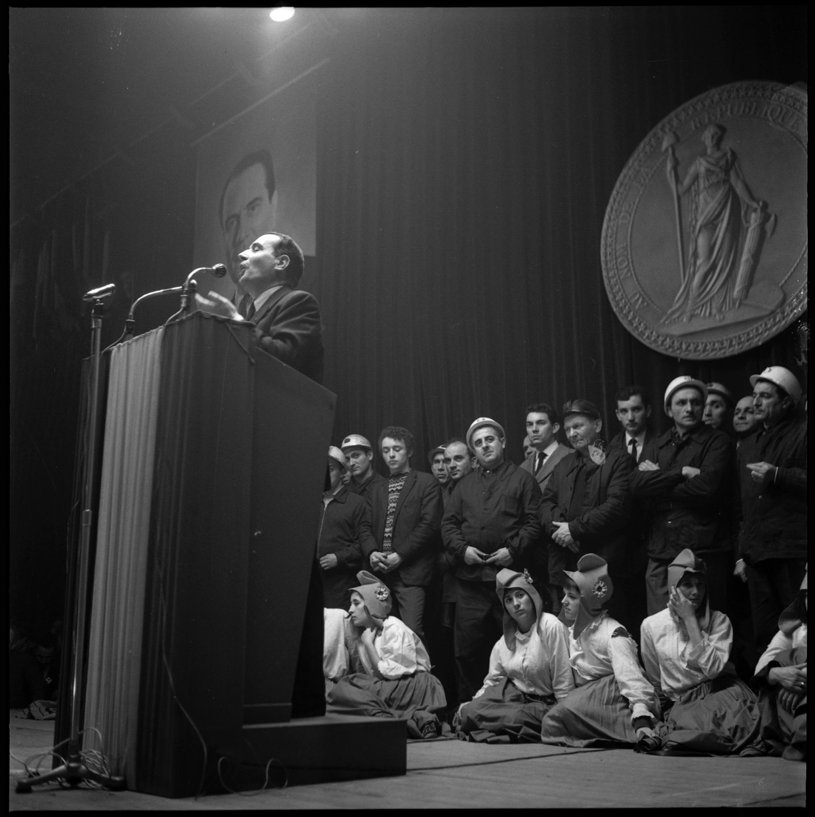 Photographie en noir et blanc, au format carré, prise du bas d'une scène éclairée par un projecteur. À gauche, François Mitterrand parle debout derrière un haut pupitre drapé, devant deux micros, la main levée. Derrière lui, sur un rideau sombre, est accroché un grand portrait photographique d'homme, celui du candidat selon toute vraisemblance. À droite, en hauteur, un très grand médaillon en relief représente une figure féminine debout, en toge, tenant une pique surmontée d'un bonnet et s'appuyant sur un faisceau de licteur ; on lit sur son pourtour « AU NOM DE LA RÉPUBLIQUE ». Sur la scène, une rangée d'hommes debout écoute, plusieurs en veste de travail et coiffés de casques de protection blancs, d'autres en costume. Au premier plan, assises par terre, des jeunes femmes en chemisier blanc et longue jupe portent un bonnet de forme phrygienne orné d'une cocarde.