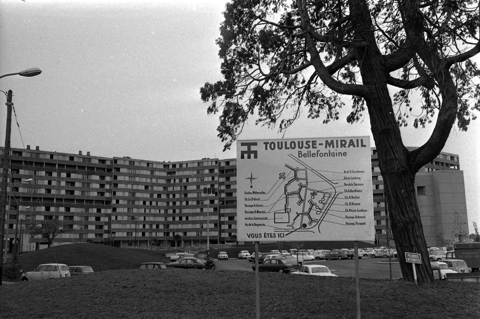 Photographie en noir et blanc, au format horizontal, prise par temps gris. Au premier plan, une terre nue et, à gauche, une butte gazonnée. À gauche et au fond, une longue barre d'immeubles d'une dizaine d'étages, aux façades rayées de bandeaux horizontaux, dont la ligne se brise en plusieurs pans. Au centre, un grand panneau blanc porte un sigle, le titre « TOULOUSE-MIRAIL Bellefontaine » et un plan du quartier : des bâtiments ramifiés en forme de Y, des voies, une rose des vents, et de part et d'autre des noms de voies et d'équipements reliés au plan par des traits ; en bas, « VOUS ÊTES ICI ». À droite, un grand arbre au tronc épais, avec à son pied une petite plaque « PELOUSES INTERDITES ». Derrière l'arbre, un immeuble plus massif et un vaste parc de stationnement rempli de voitures ; quelques voitures stationnent aussi au premier plan, et un camion bâché à droite. Des lampadaires à gauche. Aucun habitant n'est visible.